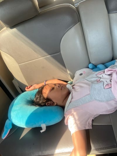 A young child is asleep in the back seat of a vehicle, resting on a blue whale-shaped neck pillow. The child is wearing a pink and grey outfit with a star pattern on it. A white plush toy with pink accents is also visible next to the child. The interior of the car appears to be made of grey leather. Sunlight is casting shadows on the car seats. The location is Bangsal, Indonesia.