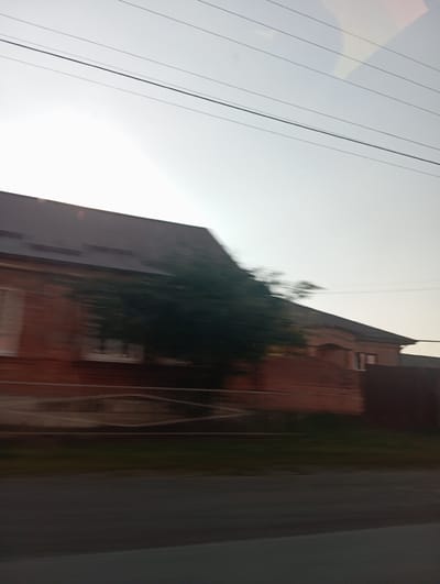 This is a side-view, motion-blurred photograph of a brick building with a dark roof, seen from a moving vehicle. Power lines are visible in the sky above the building. A fence runs in front of the building, and some greenery is present behind it. The scene appears to be in a residential area with a road in the foreground. The specific location in Gizel, Russia, is not identifiable from the visual information provided.