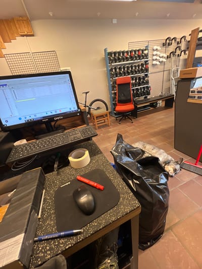 The image depicts an indoor scene within a facility in La Thuile, Italy, likely a ski and bike rental shop. The floor is covered with reddish-brown tiles, and the walls are light-colored.

In the foreground, a black speckled countertop desk on the left features a black Philips computer monitor displaying a spreadsheet application. A black computer keyboard is positioned in front of the monitor. To its right, a roll of masking tape, a black mousepad with "Fellowes" visible, and a black computer mouse with "Hemis" visible are arranged on the desk. A red marker lies near the mouse. A blue pen with "rent and go" printed on it is partially visible on the lower left of the desk. A black, tape-wrapped object rests on the far left edge of the desk.

To the right of the desk, a full, crinkled black trash bag stands on the floor. Adjacent to the trash bag, a light-colored dog with short, mottled fur and a collar lies on the tiled floor, appearing to be asleep or resting. A red-handled broom leans against a dark counter in the background, near the dog.

In the midground, a red office chair with a black backrest is placed in front of a blue and grey shelving unit displaying multiple rows of black ski or snowboard boots. To the right of the shelving unit, a dark-colored bench with a lighter top is visible. Behind the bench, a metal grid panel with white fixtures is mounted on the wall. On the far right, part of a brown counter or display unit is visible, with a "COMMENCAL" branded box on top. To the left of the boot shelving, a dark mountain bicycle is partially visible, leaning against the wall, with its rear wheel and part of its frame and seat showing. A small, light-brown wooden stool is positioned near the bicycle.

The background shows light-colored walls. On the upper left, two light-colored wooden cabinet doors are mounted high on the wall, with two white wire grid panels directly below them. No people are present in the scene.