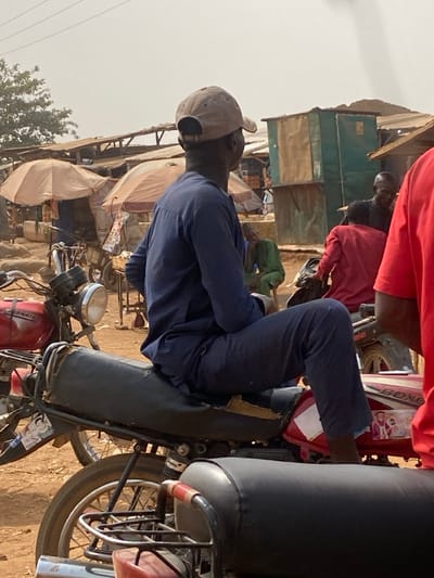 The image captures a bustling outdoor market environment in Nigeria. In the foreground, a male individual, viewed from behind, is seated on the black, torn seat of a red motorcycle. He wears a navy blue long-sleeved tunic and matching trousers, along with a beige baseball cap worn backward. His bare feet are visible. The red motorcycle features a "BOXER" decal on its fuel tank, which also bears a small sticker depicting two people.
To the left, parts of another red motorcycle with a chrome headlight and handlebars are visible. The background reveals numerous makeshift market stalls with various covers, including light-colored parasol umbrellas and corrugated metal roofs. Several individuals are present within the market: one man in a green garment is seated on a low stool in the mid-ground, while another person in a red shirt is partially visible on the far right. Indistinct figures navigate the market space. A green-painted kiosk-like structure is visible among the stalls. The ground consists of dry earth or sand, and the sky is hazy.