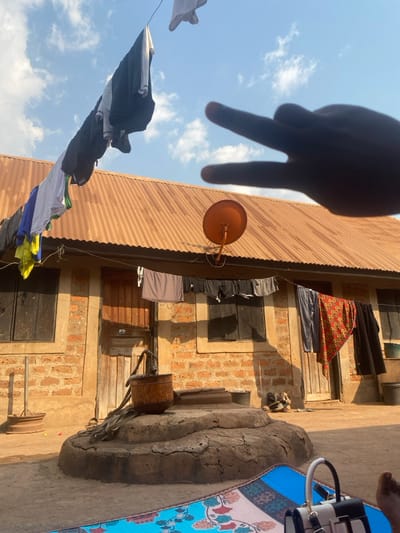 The scene is an outdoor residential area in Aliade, Nigeria, illuminated by direct sunlight under a blue sky with scattered white cumulus clouds. A blurred human right hand, with the index and middle fingers extended in a "peace sign" or "two" gesture, occupies the upper right foreground.
In the lower foreground, a blue, red, and white patterned mat lies on the ground. A black and white structured handbag rests on the mat, adjacent to a bare human foot visible in the bottom right corner.
The midground is dominated by a concrete hand-pump well, featuring a multi-tiered circular base and a rusted metal bucket attached by a rope to the pump mechanism. Immediately behind the well is a single-story building constructed with exposed red-brown bricks and topped with a corrugated metallic roof, weathered to an orange-brown color. The building's facade includes two dark wooden doors and at least two windows fitted with closed dark shutters. An orange parabolic satellite dish is mounted on the wall, below the roofline, to the right of the central wooden door.
Several clotheslines are visible. A high line stretches across the upper portion of the frame, laden with various dark and light garments drying against the sky. A lower clothesline runs horizontally across the building's facade, displaying items such as shirts, shorts, and a prominent red-and-yellow patterned fabric. Further objects include a cylindrical metal container near the left side of the building and a metal basin or bucket positioned near the rightmost window.