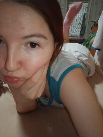 A young female with reddish-brown hair and fair skin is depicted in a close-up selfie, lying prone on a light-colored surface within an indoor residential setting in Старый Шагирт, Russia. Her head is angled towards the camera, with a visible pout and her left hand supporting her chin. She has brown eyes and a small blemish on her forehead. She is wearing a grey top with teal trim and light-colored shorts. A pink sock is visible on her elevated right foot in the upper right quadrant of the frame. The background reveals a window with textured coverings, a dark garment hanging nearby, and a partially visible white garment with blue trim to the far right.