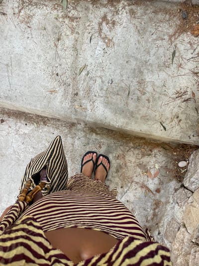 An overhead perspective shot depicts one individual standing on a light-colored, uneven concrete or stone pathway in Cap-d'Ail, France. The ground is sparsely covered with fallen pine needles and dry leaves. A horizontal step or ledge bisects the lower third of the frame. The individual wears black flip-flops and a two-piece garment or dress with an exposed midriff, featuring a maroon and pale yellow-beige horizontal stripe pattern. A woven tote bag with vertical black and tan stripes and bamboo-style handles is held on the individual's left side. To the lower right, rough-hewn light-colored stones are visible, accompanying debris and a small, smooth white pebble.