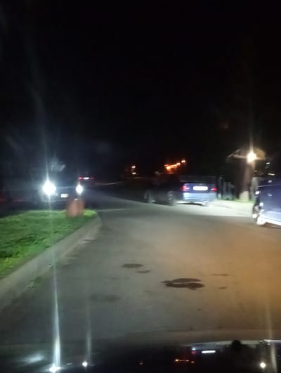 At night, a dark asphalt road curves to the right, bordered by a strip of green grass on the left. Several cars are parked along the road. In the foreground, the headlights of a vehicle illuminate the scene, creating bright streaks of light. Further down the road, a blue car with its taillights illuminated is visible. In the background, there are structures that appear to be buildings, some with illuminated windows or lights. The overall atmosphere is dark and dimly lit by artificial sources. The location is identified as Kadinski Sel'ski Sovet, Belarus.