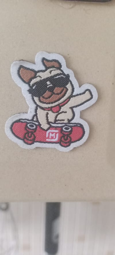 A fabric patch depicts a cartoon pug wearing sunglasses and a red collar, riding a red skateboard. The pug has its tongue sticking out and its right arm raised. The skateboard has four wheels and a white emblem with the letter "M" inside a square. The patch has a white border and is placed on a light brown, textured surface. The background is out of focus, showing vertical lines. The location in Kudymkar, Russia, is not discernible from the image.