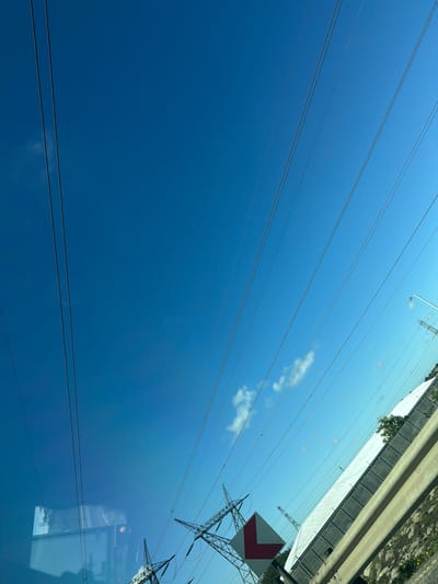The image shows a section of a road in Goodwood, South Africa with overhead power lines crossing the sky. A concrete barrier runs along the edge of the road. A road sign with a red and white arrow is attached to a power line tower. The sky is blue with a few scattered clouds. The image is taken from a low angle, looking up.