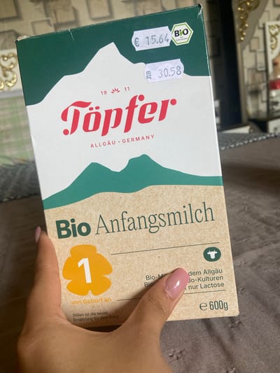 An indoor, eye-level shot captures a person holding a box of "Töpfer Bio Anfangsmilch 1" infant formula in Topoli, Bulgaria. The box, made of what appears to be cardboard with a kraft paper-like texture on part of it, is tilted at an angle, held by a hand with light pink nail polish.

The front of the box features a green and white design, typical of organic products, and clearly displays the brand name "Töpfer" in red stylized letters, with "ALLGÄU • GERMANY" indicating its origin. Prominently visible is the product name "Bio Anfangsmilch 1" (Organic Infant Milk 1), intended "von Geburt an" (from birth). A statement at the bottom reads, "Stillen ist die beste Ernährung für dein Baby" (Breastfeeding is the best nutrition for your baby).

Two price stickers are attached to the upper left corner: one showing "€ 15.64" and another, placed upside down, indicating "30.58" (without a currency symbol). A "BIO" logo with "nach EG-Öko-Verordnung" (according to EC Organic Regulation) is also visible. Other text includes "dem Allgäu", "Bio-Kulturen", and "nur Lactose". The net weight is specified as "e 600g". The box shows some minor creasing on its top left edge. The background is a blurry, patterned surface, possibly a decorative table or piece of furniture.