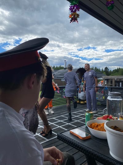 The image depicts an outdoor gathering on a wooden deck, under a cloudy sky. In the foreground, a person wearing a white shirt is partially visible, looking down at food. To the right, a black table holds a phone, a can of beer, a bowl of orange pasta, a bowl with snacks, and sliced watermelon. A large glass container filled with liquid is also present on the table.
Further back, two individuals in striped black and white prison-like outfits are interacting. One, a male, stands facing the other, who is a female with short grey hair. Behind them, other people are visible, one in an orange outfit, and another wearing a black dress and high heels. Decorative streamers are hanging from the roof structure. The background shows trees and buildings, suggesting a semi-rural or suburban environment. The overall atmosphere appears to be a casual party or event. The cropped hints do not provide specific information about the location in Joensuu, Finland.