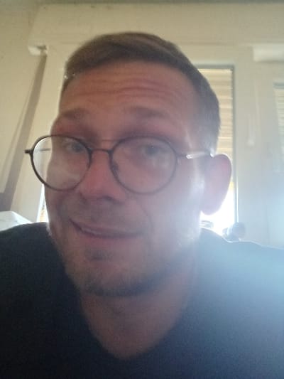 The image shows a Caucasian man wearing a dark blue t-shirt and black-rimmed glasses. He has a goatee and appears to be smiling. He is looking directly at the camera and is likely taking a selfie, evidenced by the position of the phone and the fact that the background is out of focus. The image is likely taken indoors, as can be seen from the light. The location, based on the coordinates provided, is in Montabaur, Germany, likely within a residential building.