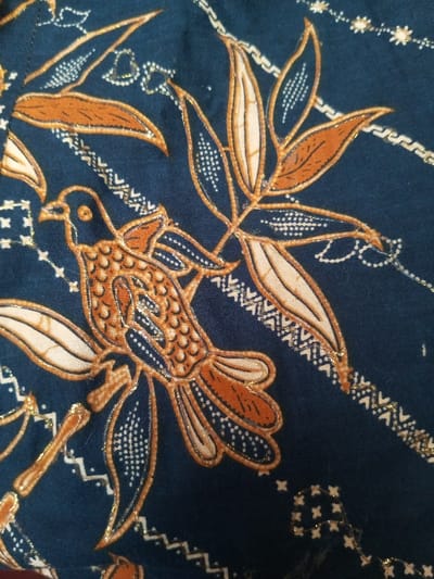 Dark blue fabric displays a repeating pattern of stylized birds and leaves. The bird is rendered in shades of brown and beige with intricate scale-like markings. The leaves are long and pointed, colored in beige and brown. Both the bird and leaves are outlined in a shimmering gold thread. Diagonal rows of geometric patterns run across the fabric, creating a textured and visually appealing design.