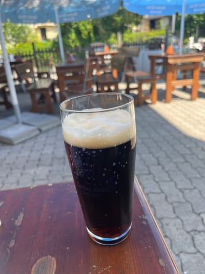 The image depicts a close-up view of a dark beer in a glass, sitting on a weathered wooden table outdoors. The beer has a frothy head and some bubbles clinging to the sides of the glass. The glass has text that reads "Becher Bräu Bayreuth," indicating the beer is from the Becher Brewery in Bayreuth, Germany. In the background, there are blurred elements of an outdoor seating area, likely a beer garden or restaurant, with wooden tables, chairs, and umbrellas. The ground is paved with interlocking stones. Sunlight casts shadows on the ground, suggesting it is daytime. There are no people clearly visible in the frame, and no specific action or interaction is depicted, other than the presence of the beer on the table.