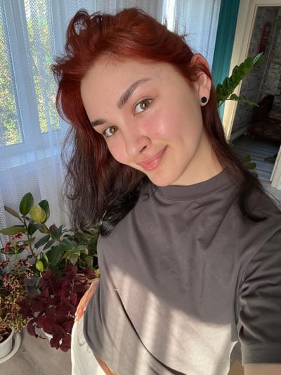 The image shows a young woman with reddish-brown hair and light brown eyes, taking a selfie indoors. She is wearing a grey t-shirt and white pants. She has a black stud earring in her left ear. Behind her, there is a window with sheer white curtains, letting in natural light. To her right, there are houseplants, including a ficus tree and a plant with dark red leaves. In the background, a wall with a textured finish and a glimpse of furniture are visible. The woman is smiling slightly, with her head tilted. The scene appears to be in a residential setting. There is no information to suggest the location is in Starobaltachevo, Russia.