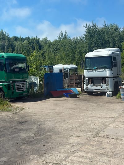 In the city of Baltiysk, Russia, an outdoor scene depicts two semi-trailer trucks, one green and one white, parked on a paved lot. The green truck, a Renault, is positioned on the left side of the frame, while the white truck is on the right. Both trucks appear to be either parked or in a state of disrepair. In the background, a dense line of trees forms a natural boundary. Scattered around the trucks are various objects, including what appear to be plastic containers and what might be spare parts for the vehicles. The ground is a mixture of asphalt and dirt, with some patches of grass. The sky above is blue with scattered white clouds, indicating a clear day.