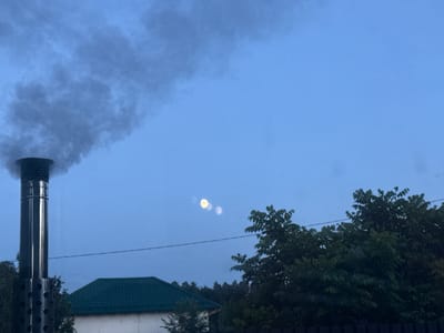 In Beryozovsky, Russia, a metallic chimney emits a thick plume of dark smoke against a clear, pale blue twilight sky. Below the smoke, the moon appears as a bright, slightly blurry orb accompanied by two fainter, circular apparitions. A dark, green-roofed structure, possibly a house, is partially visible in the lower left, with trees and foliage encroaching on the scene from the right and background. A thin, dark horizontal line, likely a wire, stretches across the frame above the building and foliage. The overall scene depicts a suburban or rural setting at dusk.