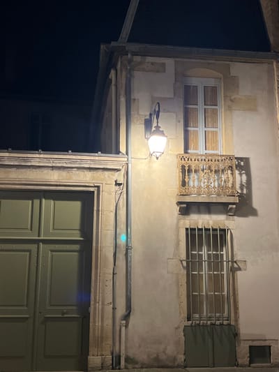 A building facade at night. A lit street lamp illuminates a gray stone wall with two windows on top, one with decorative metalwork, and a green wooden door below. A metal pipe runs horizontally above the door. The left side of the building is in shadow, contrasted with the light cast on the wall. Architectural details and textures are visible.