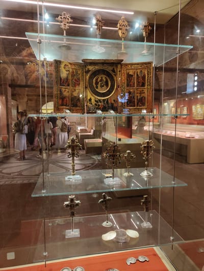 The image depicts an interior view of a museum exhibition, likely within the Holy Monastery of Great Meteoron in Kalambaka, Greece, given the religious artifacts. The scene is dominated by glass display cases showcasing religious artifacts, primarily ornate crosses of varying sizes and materials.
On the top shelf of the central display, there are several elaborate golden crosses. Below this, a triptych or polyptych painting, featuring religious scenes and figures, is prominently displayed.
The middle shelf holds several smaller, intricate crosses displayed on individual stands, some appearing to be made of metal and possibly encrusted with jewels or small decorative elements. To the right, more crosses are visible on stands, as well as what appear to be reliquaries or other small religious objects.
The bottom shelf features more crosses, again on stands, and a collection of what appear to be large, flat pearls or mother-of-pearl discs within a clear container. Further down, on an orange-red surface, there are two small, silver-toned objects that resemble antique fasteners or decorative elements.
In the background, beyond the display cases, a group of visitors, including women and men, are observing the exhibits. Their attire suggests casual wear, and some are carrying bags or backpacks. The museum environment features stone walls and architectural elements consistent with a historical monastic setting. The lighting is soft and diffused, illuminating the artifacts. Reflections on the glass surfaces indicate the presence of overhead lighting and the surrounding environment.