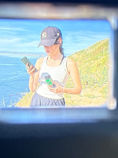 A young female individual is depicted standing on a grassy hillside overlooking the Pacific Ocean in Palos Verdes Estates, United States. The subject wears a dark blue New York Yankees baseball cap, a white sleeveless tank top, and dark shorts. A thin necklace with a small pendant is visible. She holds a dark green smartphone with a circular grip in her left hand, looking down at its screen. In her right hand, she holds a compact, dark-colored electronic device featuring a prominent green label. The background includes a vast blue ocean meeting a clear sky, with a distant landmass visible on the horizon to the left. The immediate right background shows a sun-drenched, dry-looking grassy slope. The image is framed by a dark, rectangular border, suggesting it was captured through a device's viewfinder.