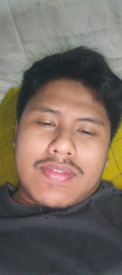 This is a close-up, overhead shot of a person lying down, facing the camera. The person has dark hair and a mustache, and their eyes are partially closed. They are wearing a dark t-shirt. The background consists of a textured surface, possibly a pillow or blanket, with a yellow checkered pattern visible on the left side. The lighting appears to be indoor, and there are no discernible external environmental cues. The overall mood is relaxed, perhaps indicating the person is resting or just waking up. The image appears to have been taken in Kemuning, Indonesia, though no specific location markers are visible within the frame. No text is present in the image.