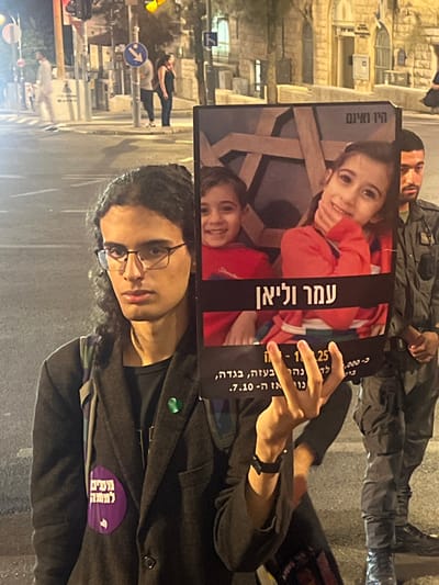 The image displays a nighttime urban scene on a major street in West Jerusalem, Israel, identifiable by its characteristic light-colored stone architecture on buildings in the background. A young individual, appearing to be male, with long dark curly hair and glasses, is positioned centrally in the foreground. He wears a dark jacket over a black t-shirt, a backpack, and a small green ribbon pin on his lapel. A purple circular sticker with Hebrew text is affixed to his jacket. He holds a large, rectangular protest sign featuring color photographs of two smiling children, a girl in a red top and a boy in a red shirt. Prominently displayed below their images is Hebrew text reading "עמר וליאן" (Omar and Liyan). Additional Hebrew text at the bottom of the sign includes the date "7.10" (October 7th) and words such as "בטוחה, בגדה". The poster's background shows a dark, abstract pattern resembling fractured wood or metal. The individual holds the sign with his right hand and forms a "V" sign with his left hand, looking directly at the camera with a neutral expression. To his right, partially visible behind the sign, stands a man in a military-style uniform, including camouflage pants and dark boots. In the background, to the left, several pedestrians are visible on a sidewalk near a pole displaying a blue directional arrow sign and a traffic light showing a red signal. The scene is illuminated by artificial streetlights.