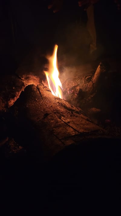 This media file captures a close-up of a campfire scene at night, likely in Khir Ganga, India. The primary subjects are the bright, flickering flames and several large logs that are burning. The fire itself is a vibrant mix of yellow, orange, and hints of blue, indicating intense heat. Wisps of smoke are visible, illuminated by the firelight, creating a hazy atmosphere around the flames and logs.

On the left side of the frame, parts of human hands are discernible, reaching towards the warmth of the fire. Their presence suggests people are gathered around, perhaps warming themselves or enjoying the ambiance. The logs forming the fire are substantial, with visible texture and charring from the heat. One large log dominates the right side of the frame, showing deep cracks and a rough surface, glowing with residual heat and embers in places. The background is completely dark, emphasizing the fire as the sole source of light in the scene.

The image depicts an event taking place at night, as indicated by the profound darkness surrounding the fire. The activity centers around the warmth and light provided by the campfire. There are no specific weather cues, but a fire often implies cool temperatures. The scene conveys a sense of primitive comfort and warmth amidst the darkness of an outdoor setting. No visible text is present in the image.