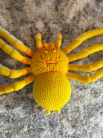 A close-up, overhead shot shows a yellow toy spider resting on a grey, textured surface that resembles a carpet or fuzzy blanket. The spider is predominantly yellow with orange accents around its head and the joints of its eight legs. Each leg is segmented, with white tips. The spider's body is round and plump, with a textured, dot-like pattern covering its abdomen. Its head features two black eyes and a red, line-like mouth. The surface it is on has a mottled grey appearance with black and white fibers. There are no discernible human elements or any indicators placing this image in Ridabu, Norway.
