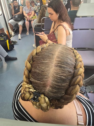 This media file is a vertical, close-up shot taken from above, focusing on the top of a person's head and their braided hair. The person is likely on public transportation, possibly a train or bus, indicated by the rows of seats with blue upholstery visible in the background and the general interior of a vehicle. The flooring appears to be a light gray.
The main subject is a woman with long, brown hair styled in a thick braid that encircles her head. The braid has a layered, textured appearance, with lighter streaks of color woven throughout. A black hair clip is visible, holding the end of the braid. Her scalp and the roots of her hair are also visible, showing a clear part down the middle.
In the background, other passengers are present, though they are out of focus. One woman to the left is wearing a white top with thin straps and a burgundy purse. Another woman in the mid-ground is wearing a patterned top and shorts. A man in the lower left is wearing a black t-shirt and has tattoos on his arm. Another person in the lower right is wearing black shoes and grey pants.
The lighting suggests it is daytime, likely indoors. There are no strong indicators of the specific time of day or weather conditions. The overall mood is casual and everyday, with no discernible strong emotions displayed by the visible individuals.
There is no visible text within the image. The overall scene is a snapshot of life on public transport, with the focus primarily on the hairstyle.