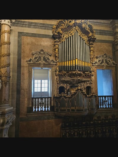 This is a medium shot of a large, ornate pipe organ mounted on a wall within a building. The organ has numerous metallic pipes of varying lengths, some with gold accents at the top. It is surrounded by intricate gold-colored decorative carvings. To the left of the organ is a tall, fluted column with gold floral decorations. On either side of the organ, there are windows with ornate wooden balconies. The window on the left has a closed, white blind, while the window on the right shows a bright blue, likely exterior view. The walls appear to be made of stone with marbled patterns. The lighting is dim, with the organ and its decorations being the most illuminated elements. The image does not contain any people or visible actions.