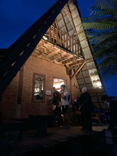 A low-angle shot captures the exterior of a two-story brick building with visible wooden frame construction under its roof, set against a dark blue twilight sky. A fern-like plant dangles in the upper left corner. Two windows are visible in the brick facade. One window has a dark metal frame with visible bars and the other window appears to have a light interior. Figures stand on a dark platform on the right side of the frame. One is wearing a white shirt and appears to be looking downward. Other dark shapes obscure the details of the platform and figures. This picture was taken in Tamiajeng, Indonesia.