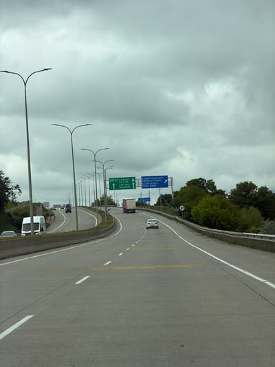 The image shows a highway in Mrgvali Chala, Georgia, under a cloudy sky. The road curves to the right and has multiple lanes. Vehicles, including cars and trucks, are present on the road. Road signs with Georgian and English text are visible, along with tall street lights lining the side of the road. Trees are present along the edge of the highway.