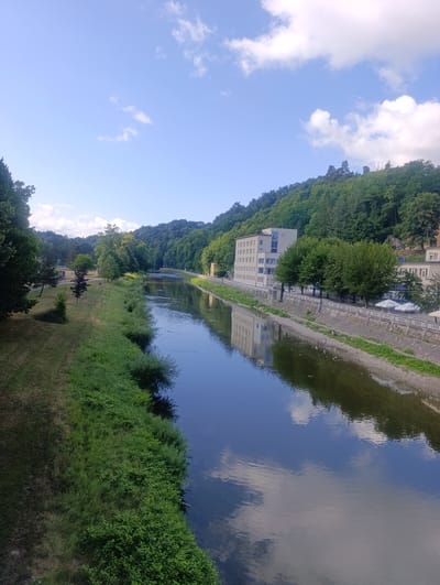The image shows a wide river flowing through a valley with a paved embankment on the right side and a grassy bank on the left. A large, light-colored building with multiple windows is situated along the embankment, reflecting in the water. Trees line the embankment in front of the building, and a stone wall runs alongside it. The left bank is covered in grass and dense vegetation, with scattered trees and a path. In the distance, the river curves towards a densely wooded hillside under a blue sky with scattered white clouds. The scene appears to be the vicinity of Teplice nad Bečvou, Czechia, given the characteristic landscape with a river and valley.