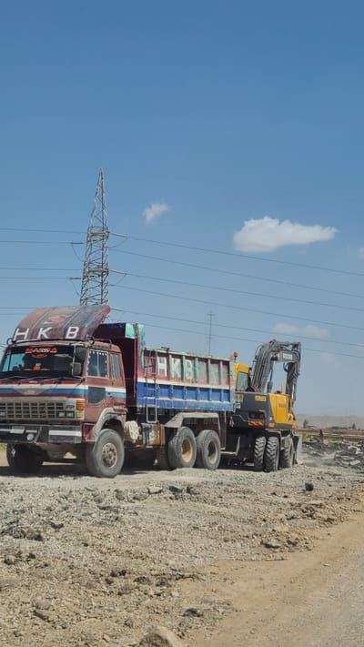 The image displays a daytime scene featuring heavy machinery and an industrial landscape, rotated 90 degrees clockwise. In the foreground, a large, well-worn red dump truck is prominently parked on rocky, dirt ground. The truck's cab has "HKB" painted in large white letters on its side, and some red text, possibly including "Hino," below it. The truck appears to be carrying a blue and green dump body, which is also weathered.
Behind and to the right of the red truck, a yellow Volvo excavator is visible. The brand "VOLVO" is clearly readable on its arm and body. The excavator is also parked on the gravelly terrain, near what appears to be a pile of construction material or rocks. The presence of the dump trucks and excavator suggests an active construction or earthmoving site.
In the background, a tall metal electricity pylon or transmission tower stands prominently, with power lines extending across the bright blue sky. The sky is clear with a few scattered white clouds, indicating sunny weather. The ground is covered in loose rocks and dirt, suggesting an arid or semi-arid environment, possibly a rural area or a construction zone outside a city. Distant hills or mountains can be seen on the horizon. The overall impression is one of heavy industry or infrastructure development in a robust, open environment.
