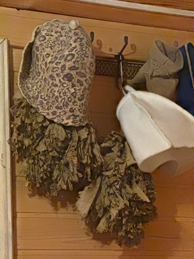 In Mayak, Russia, inside a wooden sauna, two bath whisks (venik) are hanging on a wall. One whisk has a leopard-print felt hat on top, and the other has a white felt hat. A light brown, possibly cotton, hood is also visible, along with a dark blue cloth object, hanging from hooks on the wooden wall. The whisks, made of dried branches and leaves, are positioned to the left and right, with the leopard-print hat in the center-left and the white hat in the center-right.