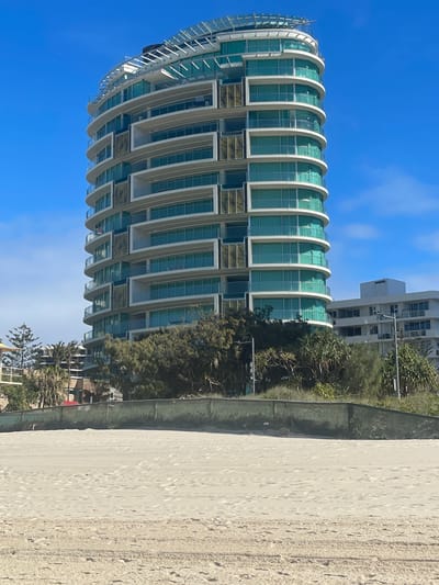 A tall, modern, circular apartment building with a glass facade and balconies stands on a beach on the Gold Coast, Australia. The building has a white frame with curved balconies and sections of glass that reflect the sky and surrounding trees. The sky is bright blue with a few wispy clouds. In front of the building is a sandy beach with tire tracks and footprints. Behind the building, there are more trees and other buildings. The overall scene is one of a sunny day at a beachside resort.