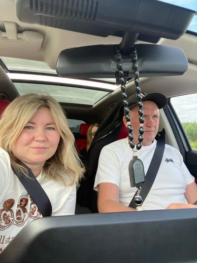 This media file is a selfie taken from inside a car, with the camera pointed towards two people. The subjects are a man and a woman. The man is in the passenger seat, wearing a black baseball cap and a white t-shirt. He has a string of black oval beads with silver accents hanging from his rearview mirror, draped over his shoulder. The woman is in the driver's seat, looking towards the camera and smiling. She has blonde hair and is wearing a white t-shirt adorned with sequined Mickey Mouse figures. Both individuals are wearing seatbelts.
The scene appears to be a road trip, likely during the daytime, given the bright lighting and the view of trees and sky through the car windows. The weather seems to be overcast. The interior of the car is visible, including the rearview mirror, dashboard, and car seats. The red upholstery of the seats is visible behind the woman.
There is some visible text on the man's t-shirt; it appears to be a logo in white. The tag on the rearview mirror also has some text and numbers, which are partially obscured. The woman's t-shirt also has some text along the bottom, but it is largely cut off by the edge of the frame. The overall mood appears to be casual and happy, suggesting a pleasant journey. The location context provided is Поварёнки, Russia.