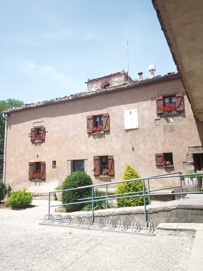The image shows the exterior of a building in Susqueda, Spain, likely a rural house or inn. The building has a light brown, textured facade with several wooden-shuttered windows, some of which are adorned with red flowers. A sundial is mounted on the wall above a smaller window. The roof is tiled with red clay tiles. In front of the building, there is a paved courtyard with cobblestones and a bicycle rack. There are also various plants and bushes, including a small, conical evergreen tree. A metal railing runs along a raised walkway or terrace. The sky is clear blue with a few wispy clouds. The scene appears to be bathed in sunlight, indicating it is daytime.