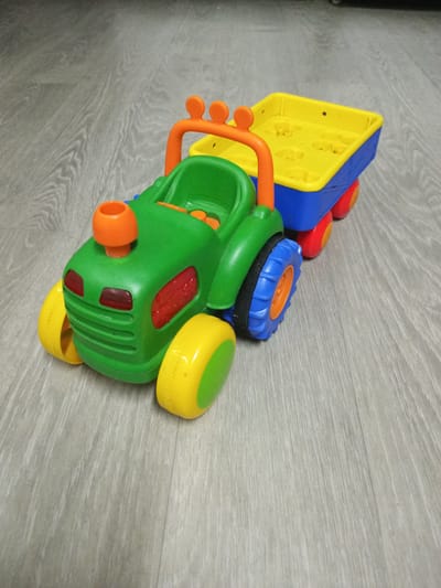This is a medium shot, eye-level, close-up photograph of a colorful toy tractor with a trailer. The toy is made of plastic and features bright green for the tractor body, a yellow trailer with blue sides, and orange accents on the tractor's exhaust pipe and roll cage. The wheels are yellow with green accents on the front and blue with orange hubs on the back. The trailer has a textured yellow surface with molded shapes, possibly representing hay bales or other cargo.
The toy is positioned on a light gray wooden floor, which appears to be made of laminate or wood planks with visible grain patterns. The lighting is even, suggesting an indoor setting, and there is no indication of time of day or weather conditions. The image is sharp and well-focused on the toy.
The scene is a simple, static shot of a toy. There are no people visible, and the activity is simply the toy resting on the floor. The location context provided is Shostka, Ukraine, but there are no specific visual cues within the image to confirm this location. No text is visible in the image.