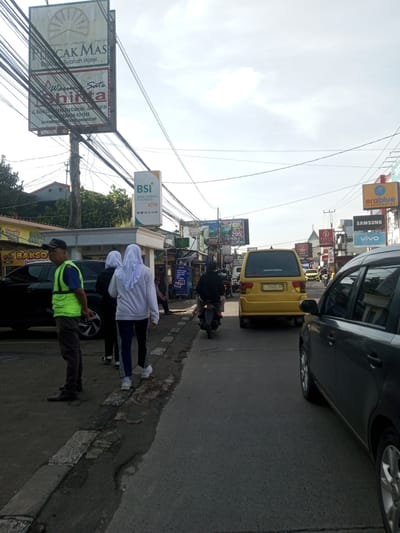 The image captures a street scene in what appears to be an urban or suburban area. There is a mix of traffic, including cars, a yellow van, and a motorcycle. Several pedestrians are present, including two women wearing headscarves and a man wearing a reflective vest standing on the sidewalk. A large sign for "PUC CAK MAS Syariah Hotel" and other businesses such as "BSI BANK SYARIAH INDONESIA" and "erablue ELECTRONICS" are visible. Various other commercial signs are scattered along the street. The environment is cluttered with wires overhead. The location is presumably somewhere with Islamic presence with the hotel sign. The license plate on the yellow van reads "S 1969 YT". The specific city is not visible within the image.