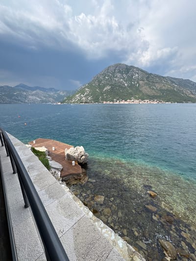 This is a high-angle, vertically oriented, wide shot of a scenic coastal view near Đurići, Montenegro. The scene features a calm, clear blue body of water, likely a bay or lake, with a rocky shoreline and a lush green, mountainous landscape in the background.
In the foreground, a stone walkway with a dark railing lines the bottom of the frame. Adjacent to this, a small, wooden platform juts out into the water, built around a large rock. The water is transparent near the shore, revealing the submerged rocks and pebbles.
Further out, the water deepens to a rich blue. Two small, orange buoys are visible floating on the surface. The mountainous terrain behind the water is covered in dense green vegetation, and a small, picturesque town with white buildings and red roofs is nestled along the coastline. The sky is partly cloudy, with dramatic grey clouds present, suggesting a dynamic weather pattern.
The overall atmosphere is serene and tranquil, highlighting the natural beauty of the Montenegrin coast. There is no visible text in the image. The lighting suggests it could be late afternoon or early evening, given the interplay of light and shadow on the mountains and the water.