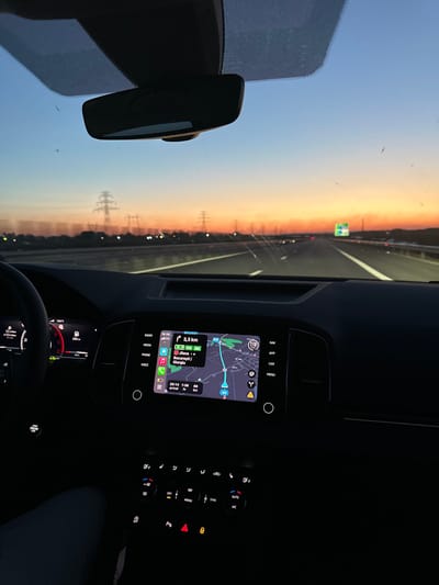 The image depicts the interior of a vehicle from a first-person perspective, looking forward through the windshield at dusk. The vehicle is traveling on a multi-lane highway under a sky transitioning from orange at the horizon to blue overhead, consistent with a sunset. Visible through the windshield are multiple electricity pylons stretching into the distance along the highway's edge, and a green road sign with white text on the right side of the road. The windshield exhibits numerous small specks, possibly dust or insects.
Inside the vehicle, a dark dashboard occupies the lower portion of the frame. A rearview mirror is mounted centrally at the top of the windshield. The left side shows a portion of the driver's steering wheel and an illuminated digital instrument cluster displaying a speedometer reading, fuel level, and other vehicle data. Centrally located on the dashboard is an infotainment screen displaying an active navigation map. The map indicates "3.3 km" remaining, with destinations "Jilava," "București," and "Giurgiu" listed, and a timestamp of "20:14." These locations place the vehicle within Romania, specifically in the vicinity of Sintești, which lies south of Bucharest on routes toward Giurgiu. Below the screen, illuminated climate controls and vehicle function buttons are visible, including the active hazard lights indicator. No individuals are directly visible, but the scene implies a driver is operating the vehicle.