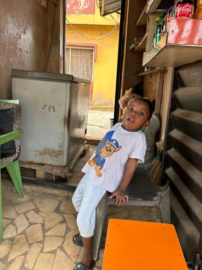 An image taken in Ashipa, Nigeria, depicts a young Black child standing within what appears to be a small shop or entryway, looking directly at the camera. The child, wearing a light grey "Paw Patrol" t-shirt, light blue cropped pants, and black Adidas slide sandals, leans with his left hand on a dark stool or counter. His head is tilted slightly, and his mouth is slightly open, revealing his tongue.

To the child's left, a large, weathered, light-grey chest freezer rests on wooden pallets. To his immediate right is a bright orange, flat surface, possibly a stool or small table. Behind the child and to the right, a dark, slatted structure holds shelves stocked with various bottled beverages, including Coca-Cola, and other items like a broom and plastic bags.

The interior space features a tiled floor with irregular light-colored stones and peeling, light-colored walls. An open doorway or passage in the background reveals an exterior urban setting. This outdoor area features a dominant yellow building with a white-framed window, showing signs of wear and peeling paint. Electrical wires are visible strung across the exterior scene. A section of a building with red-orange peeling paint and a corrugated metal roof edge is visible at the top of the background.