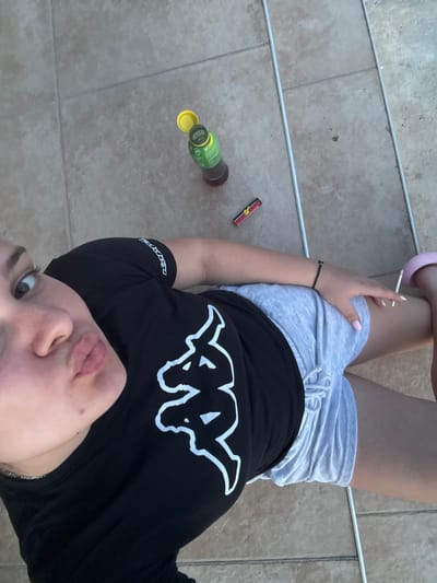 In the city of Rogosh, Bulgaria, a young woman lies on a tiled patio. She is wearing a black t-shirt with a white Kappa logo and grey shorts. She is holding a cigarette and a pink flip-flop is visible on her foot. In the background, a bottle of what appears to be iced tea and a lighter are on the ground.