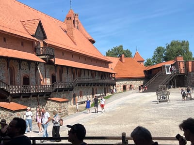 The image depicts the Trakai Island Castle in Trakai, Lithuania, on a sunny day. The castle, constructed of red brick with distinctive red-tiled roofs and wooden balconies, dominates the left side of the frame. Multiple tourists are visible walking around the castle grounds. In the foreground, several people are seated or standing, appearing to observe the scene. In the middle ground, a group of tourists walks along a path leading towards the castle. Further back, a set of wooden stairs ascends to a higher level of the castle. A historical pillory-like structure is present in the courtyard. The ground is covered in gravel and sand, and trees are visible in the background. The sky is clear and blue.