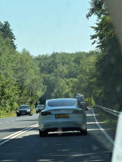 A rear-view, medium-shot captures a white Tesla Model 3 driving on a paved road through a forested area in Niederanven, Luxembourg. The Tesla is in the foreground, with its license plate, "DK 1987," clearly visible. The road has double white lines, indicating it is a two-lane road with traffic going in the same direction. A black Mercedes-Benz sedan is driving ahead of the Tesla in the left lane. Further ahead, other vehicles, including what appears to be a truck, are visible, suggesting moderate traffic. The road is flanked by dense, green trees on both sides, with a metal guardrail visible on the right. The sky is a clear, light blue. The image has been taken from another vehicle, with a portion of its windshield visible in the lower right corner.