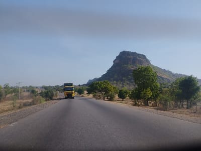 A single yellow semi-trailer truck travels on a paved road in the direction of a rocky, barren mountain in the distance. The road is flanked by scrub brush and small trees. The sky is clear and blue. This location is near the city of Ture in Nigeria, close to coordinates (latitude: 9.812, longitude: 11.381).