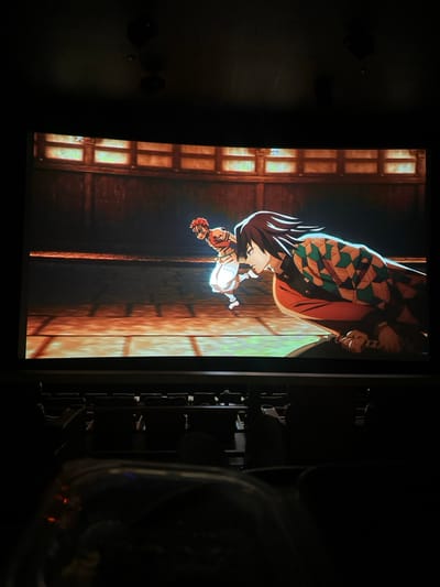 An interior view of a movie theater in Paradise, United States, depicts an animated scene projected onto a large, curved screen. The screen prominently displays two male anime characters engaged in dynamic action within a traditional wooden arena setting. The character on the left has reddish-orange spiky hair, wears a striped orange top and white pants, and is shown in mid-stride. The character on the right has dark hair, wears a distinctive patterned haori over a red garment, and is crouched low, with a sword hilt visible near his hip. The arena features a wooden floor and upper wall sections with rectangular, dimly lit panels. Below the screen, several rows of dark, empty cinema seats are visible. The foreground includes a partial view of a dark container, possibly a cup or popcorn tub. The theater ceiling is dark, with two black circular objects (likely speakers) mounted. The overall environment is dark, typical for a cinema during a screening.
