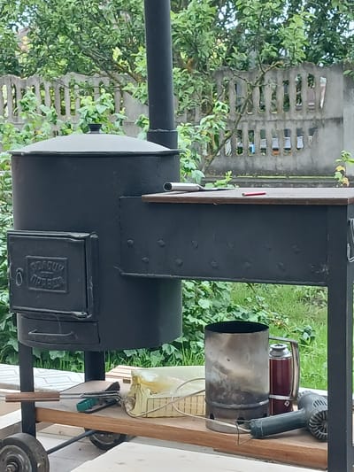 This is a medium shot of a black metal outdoor barbecue grill and smoker with a vertical chimney, situated on a wooden cart with wheels. The grill has a side-mounted work surface and a small door with what appears to be Cyrillic text embossed on it. On the work surface, there is a metal spatula and a red object, possibly a lighter. Below the work surface, on a wooden shelf attached to the cart, are several items including a metal chimney starter, a red thermos, and a dark green electric hair dryer. In the background, there is a concrete fence with rectangular openings, and lush green foliage from trees and bushes. The scene is outdoors, likely in a backyard or garden area. There are no people visible in the image.