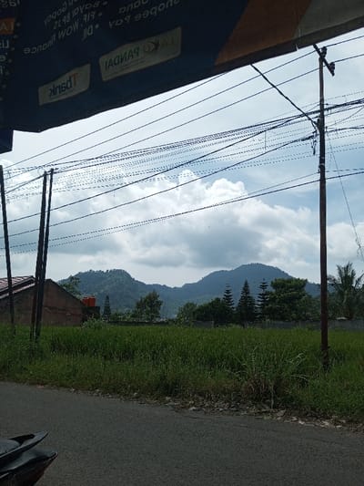 The scene is a daytime exterior in the city of Namthoung, Laos. The foreground features a dark asphalt road running from the lower left, bordered by overgrown green grass and weeds. A black motorcycle seat and rear fender are partially visible in the bottom left. Directly overhead, an inverted dark banner displays fragmented white text, including "K PANDAI" and "Bank". The midground includes multiple dark utility poles with a dense network of black overhead wires crisscrossing against a cloudy sky. A flat expanse of uniform green vegetation, consistent with an agricultural field, is present behind the foreground elements. Behind this field, on the left, a reddish-brown building with a dark roof and an orange cylindrical water tank is observed. A grey concrete wall extends horizontally across the frame behind the field. The background consists of a prominent range of lush, tree-covered mountains beneath a sky populated by significant white and grey cumulus clouds. Various trees, including several tall conical evergreens and a palm tree on the far right, are situated at the base of the mountains. No human figures or discernible activity are present.