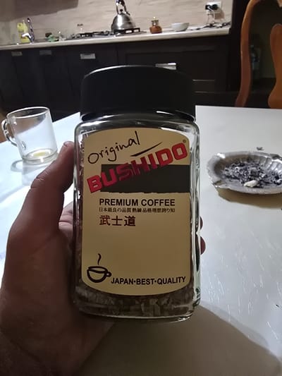A hand holds a glass jar of "Original BUSHIDO PREMIUM COFFEE" against a plain white surface, with a kitchen counter and cabinets visible in the background. The jar's label features Japanese characters and the text "JAPAN·BEST·QUALITY". To the right of the jar, an ashtray filled with cigarette butts and ash sits on the surface. The scene appears to be indoors, in what looks like a home kitchen. No specific location in Khasavyurt, Russia can be determined from the visual information provided.