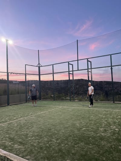 Two people are playing padel tennis outdoors in the city of El Poble Nou de Benitatxell/Benitachell, Spain. One person is wearing a white shirt, black pants, and a white hat, while the other is wearing a gray shirt and black shorts. Both are holding padel rackets. The padel court is surrounded by a metal cage and net. The background features a landscape with trees and a sky with a pink and purple sunset.
