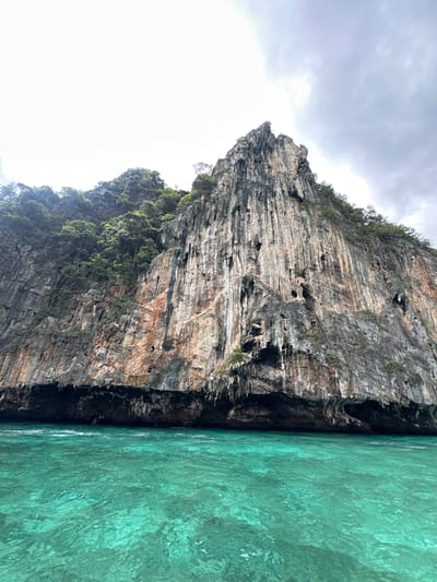 This is an eye-level, full shot of a limestone karst formation rising dramatically from the vibrant turquoise waters of Haad Yao, Thailand. The sheer cliff face is weathered and striated, with patches of orange and grey rock, and dotted with numerous small caves and crevices. Lush green vegetation clings to the upper slopes and crest of the formation, contrasting with the barren rock. The water is clear and slightly choppy, reflecting the overcast sky. The overall impression is one of natural grandeur and a remote, tropical seascape. There are no people or other objects of interest visible in the image.